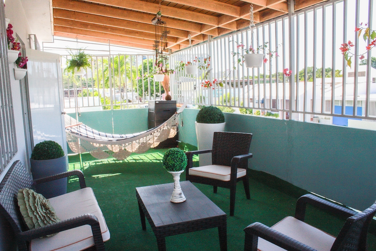 A covered balcony space is furnished with comfortable seating, including two chairs and a low table. Decorative plants are positioned around the area, complemented by a hammock for relaxation. Natural light fills the space, creating an inviting outdoor retreat.