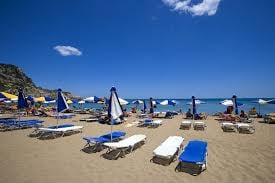 A sandy beach is depicted, featuring several rows of lounge chairs with blue and white striped umbrellas. People are scattered across the beach, relaxed under the sun, while gentle waves can be seen in the distance, accompanied by a clear blue sky with a few clouds.