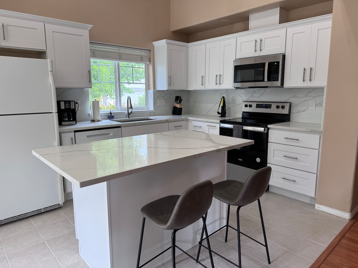 The kitchen features modern cabinetry in a light finish, complemented by a marble countertop and an island with seating for two. Stainless steel appliances, including a microwave and oven, are included. Natural light enters through the window, illuminating the spacious layout.
