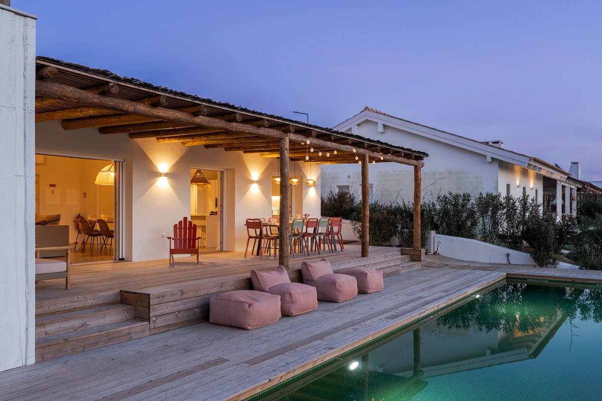 An inviting outdoor deck features comfortable seating and a dining area under warm lighting. A restful pool reflects the sky, surrounded by lush greenery. The exotic wood structure adds a rustic charm, enhancing the seamless connection to the indoor space.