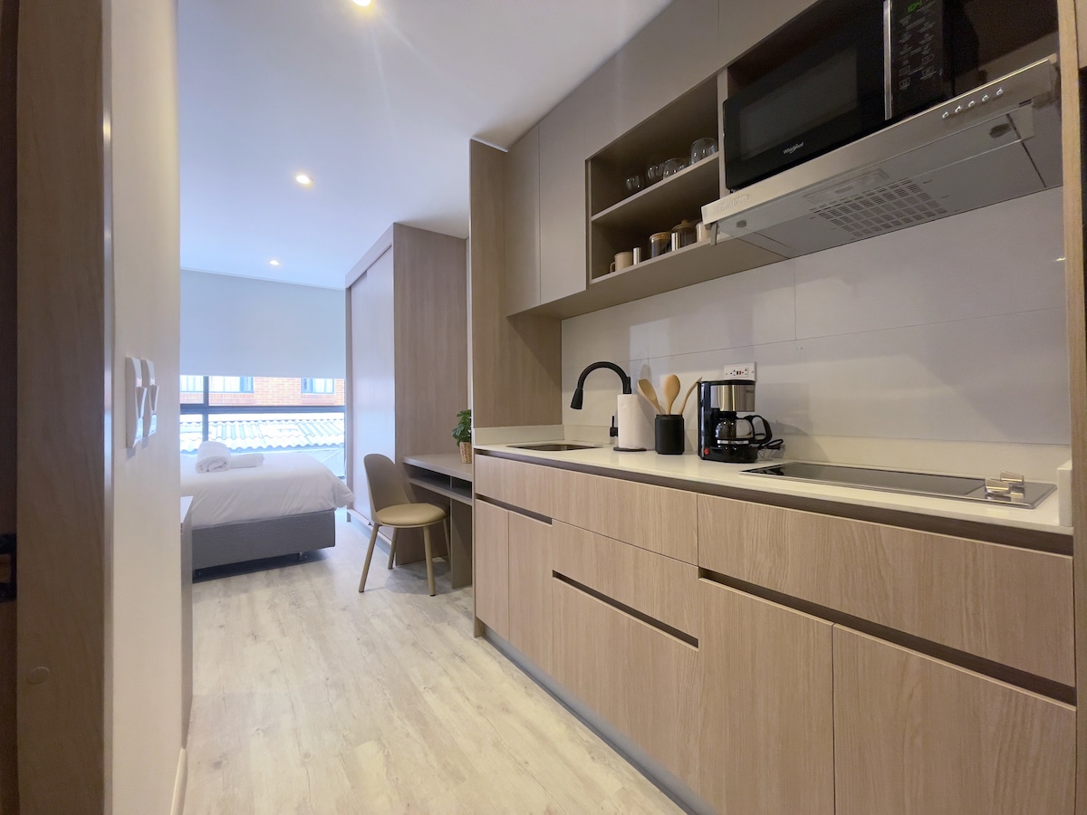 A modern kitchen area features wood cabinetry and countertop space, including essential appliances such as a refrigerator and microwave. A dining chair is positioned nearby, with natural light entering from the adjacent room, which contains a spacious bed.