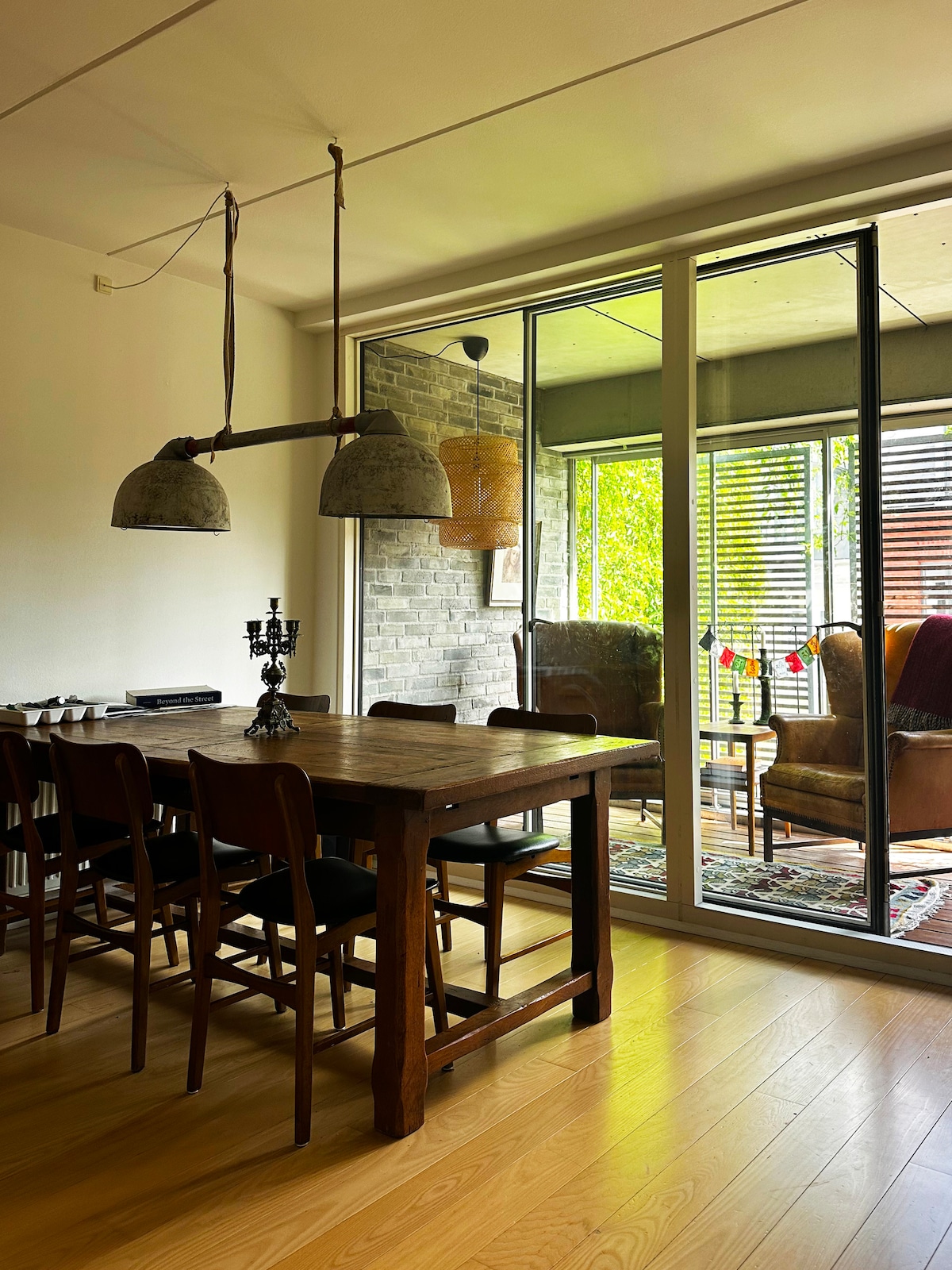 A dining area features a large wooden table surrounded by dark wooden chairs. Above, pendant lights hang from the ceiling. Floor-to-ceiling glass doors lead to a cozy terrace with greenery visible through the slats, enhancing natural light in the space.