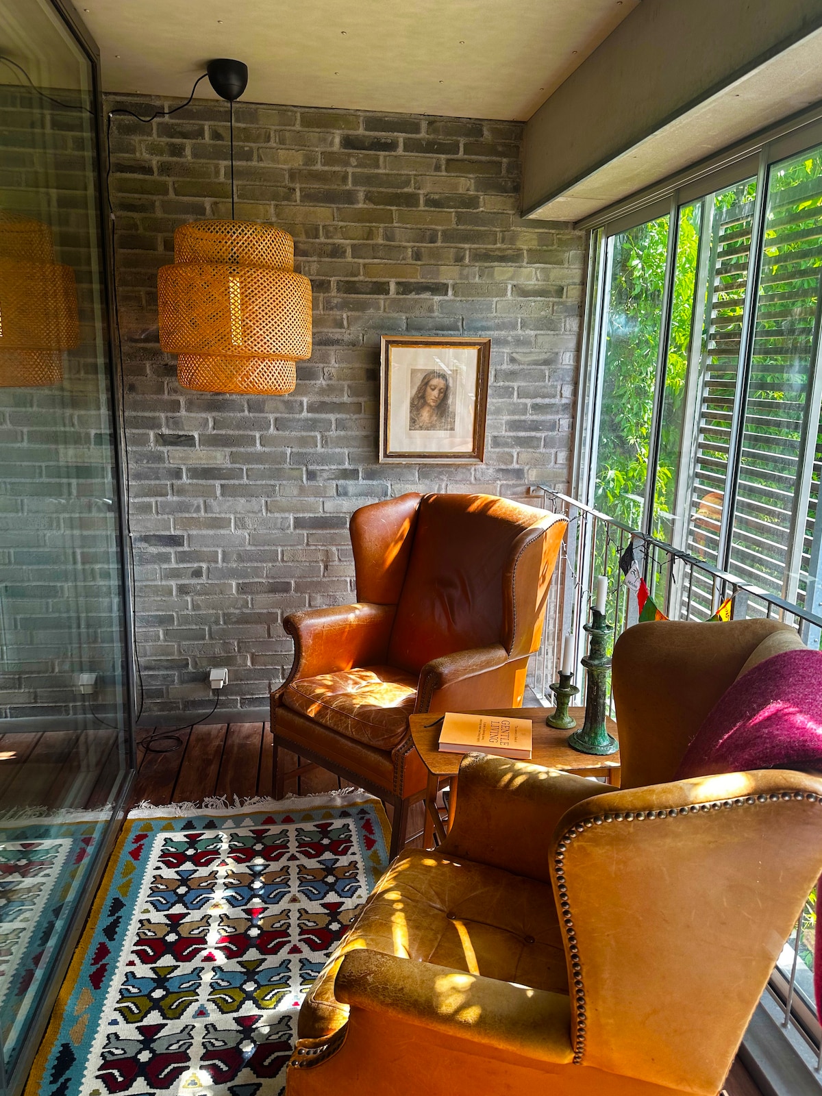 A cozy glass-covered terrace features two armchairs positioned beside a small wooden table. A vibrant area rug covers the floor, and a woven pendant light hangs overhead. Natural light filters through the glass, highlighting a piece of framed art on the brick wall.