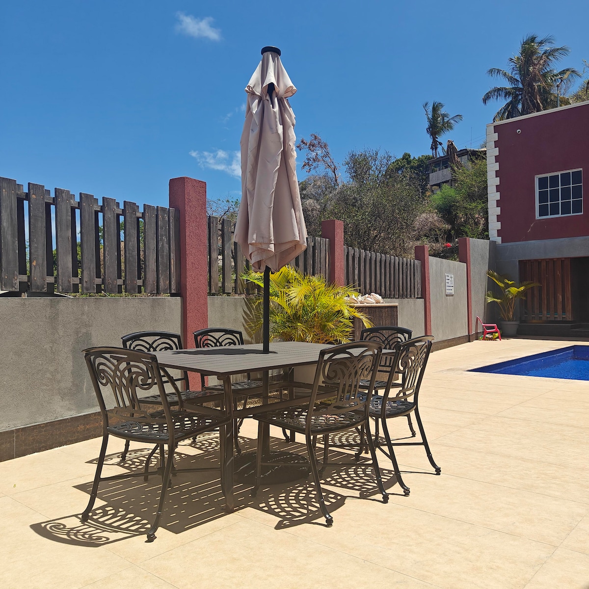 An outdoor dining area is featured, surrounded by a stone patio. Six metal chairs encircle a rectangular table, with a large umbrella positioned at the center for shade. The backdrop includes a swimming pool and tropical vegetation, set under clear blue skies.