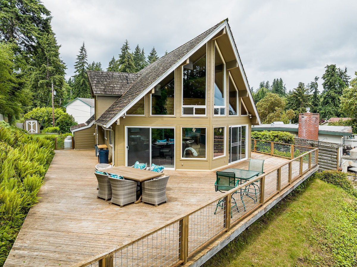 A spacious deck wraps around a modern house featuring large glass windows that highlight the open interior. The inviting outdoor space includes a dining table, comfortable seating, and lush greenery surrounding the property. The sloped roof adds architectural interest to the design.