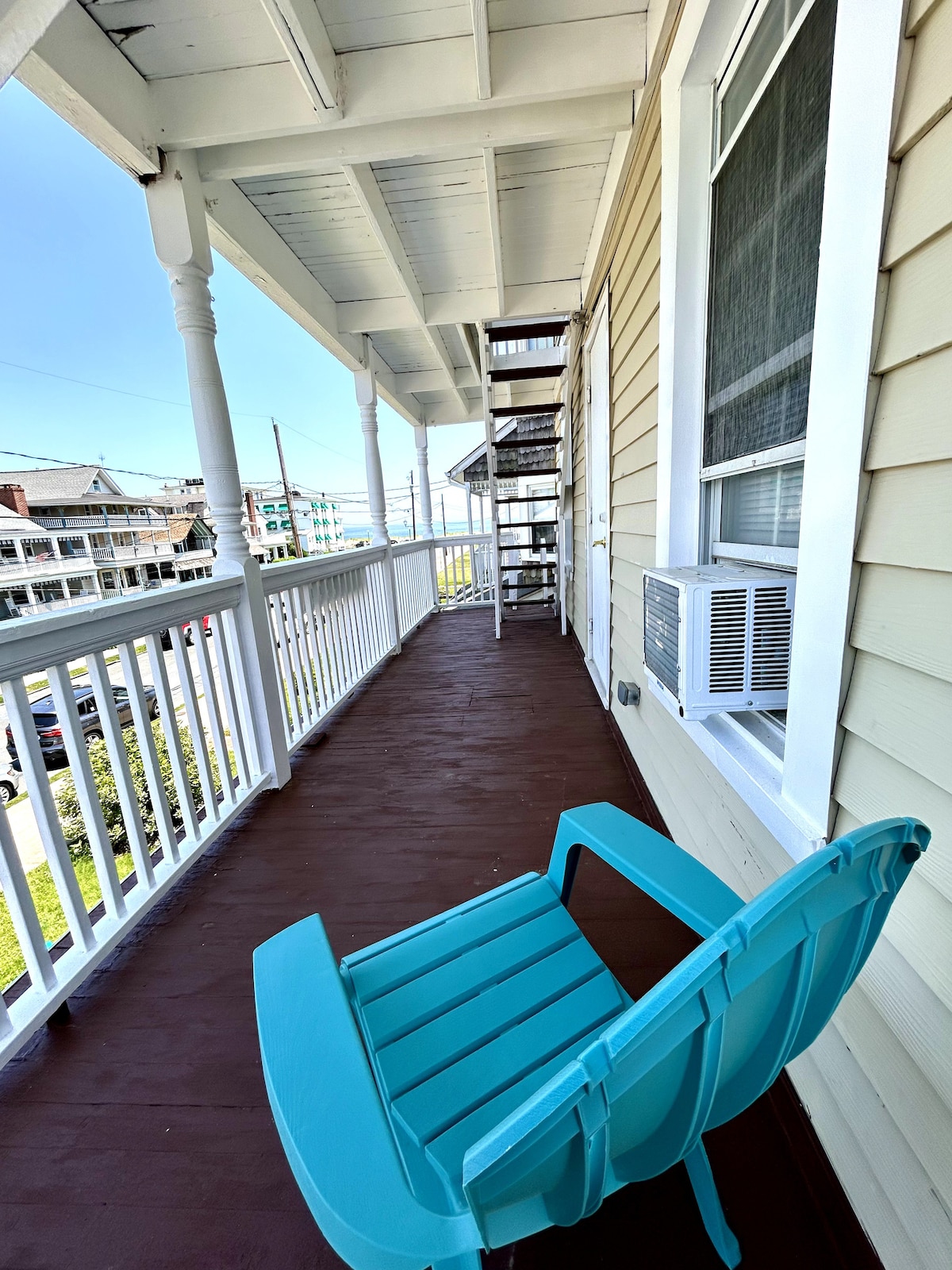 A spacious balcony is featured, lined with white railings and equipped with a turquoise chair. An air conditioning unit is mounted on the wall, and a spiral staircase is visible in the background. The view includes a sunny sky and nearby structures.
