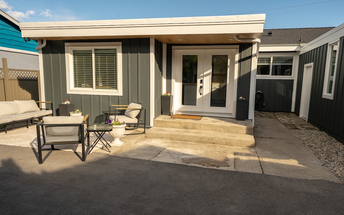 A modern entrance to a private rancher suite is shown, featuring steps leading to a double-door entryway. Comfortable outdoor seating is arranged with small tables, surrounded by potted plants. Natural light highlights the inviting exterior, which is accented by horizontal planks in muted tones.