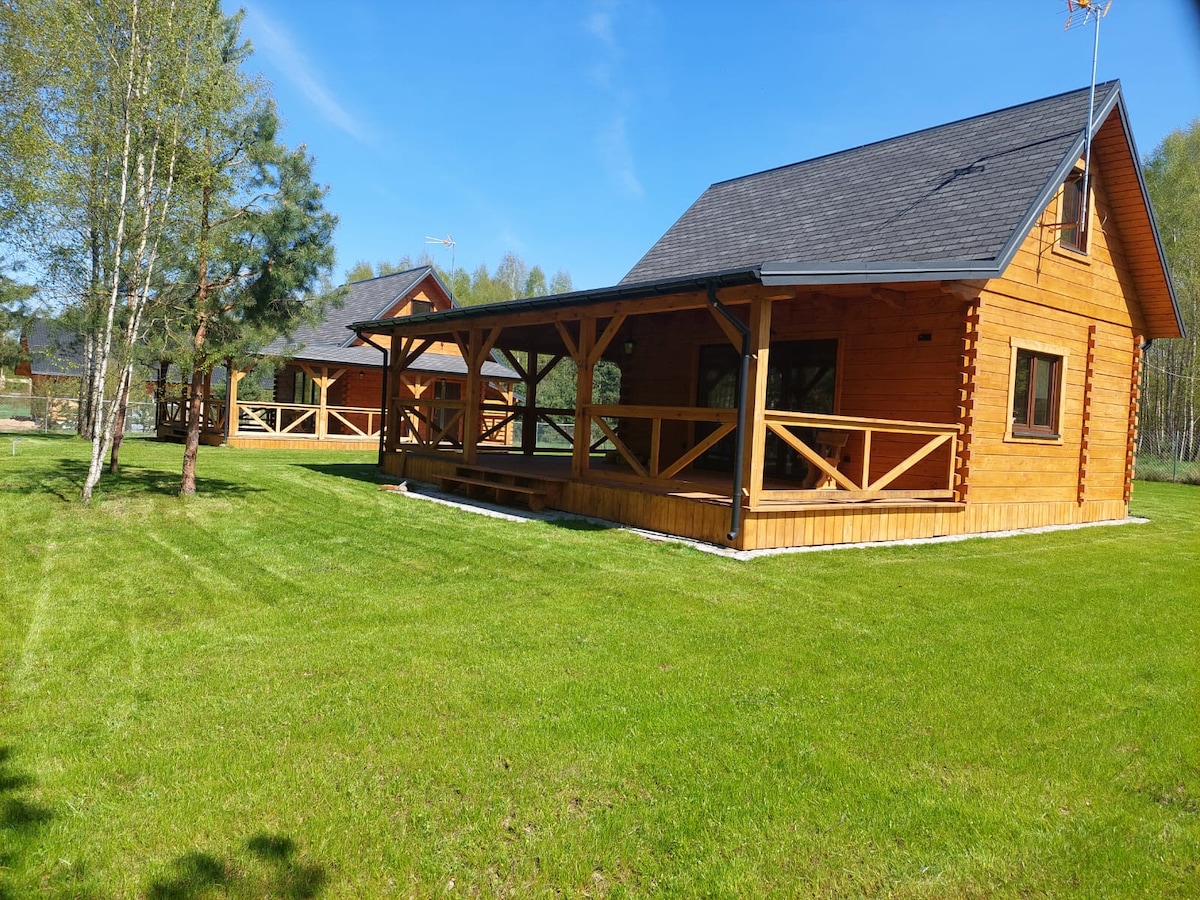 Two wooden cabins are visible on a spacious green lawn. Each cabin features a porch with wooden railings, surrounded by a few trees. The clear blue sky provides a serene backdrop, and the grassy area extends around the cabins, enhancing the outdoor setting.