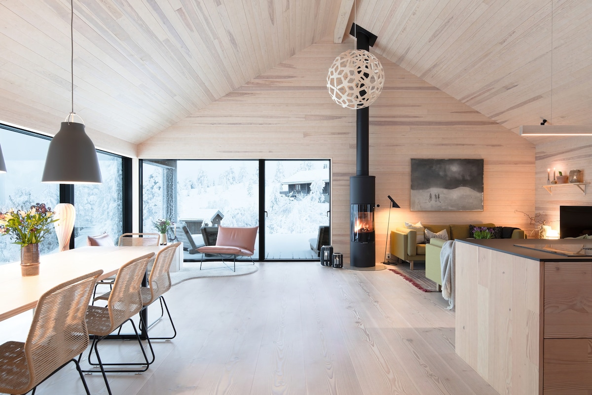 An open living space features tall ceilings and large windows that offer views of the snowy landscape. A modern dining table is surrounded by six chairs, while a cozy seating area with a fireplace and artwork creates a calming focal point.