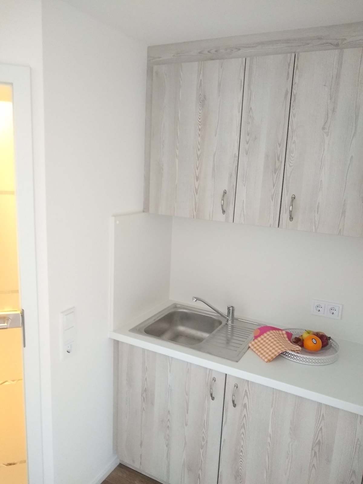 A compact kitchenette features light wood cabinetry and a stainless steel sink. A small assortment of fruits is arranged on a plate, with a decorative kitchen cloth nearby. The bright and neutral walls contribute to a clean and uncluttered appearance.