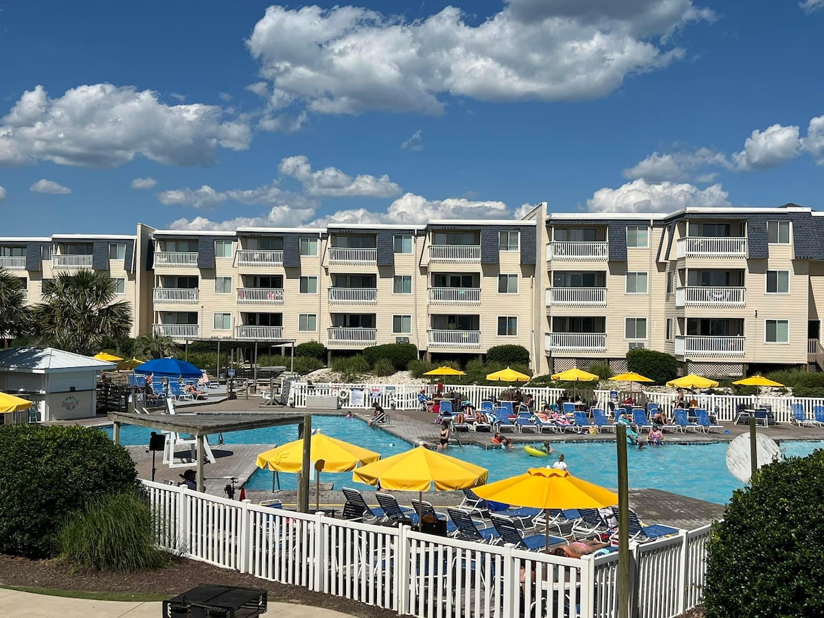 A spacious outdoor pool area is surrounded by numerous yellow umbrellas and lounge chairs. The two-story condo building in the background features multiple balconies. Green shrubs and well-maintained lawns enhance the inviting atmosphere, with a clear blue sky and clouds visible above.