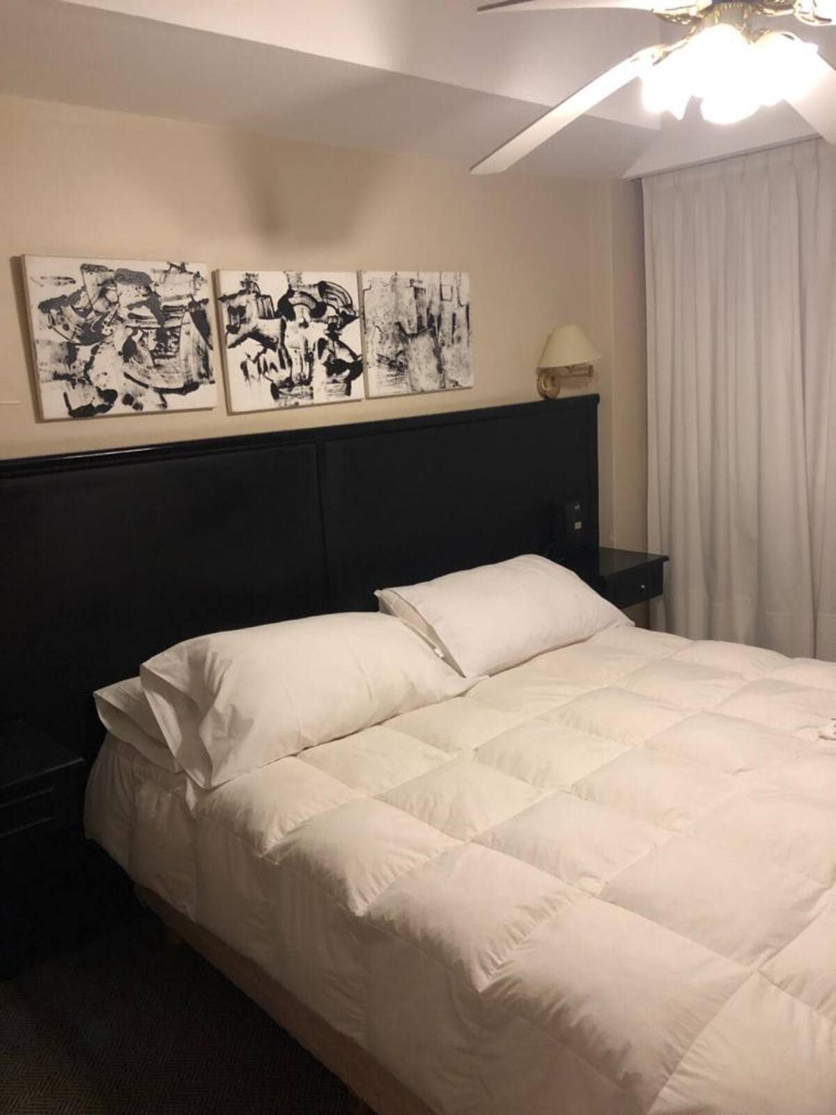 A comfortable bedroom showcases a spacious bed topped with white bedding. Black and white artwork is displayed above the headboard, while a bedside lamp is positioned on either side. Natural light filters through sheer curtains, creating a calm environment.