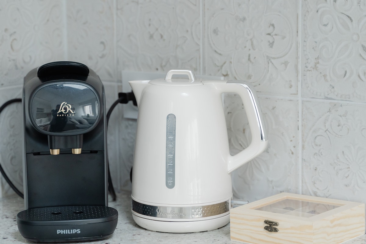 A coffee machine with a sleek design is positioned next to a stylish white electric kettle. The kettle features a clear water measurement gauge and chrome accents. A simple wooden box is also visible on the countertop, enhancing the kitchen space.