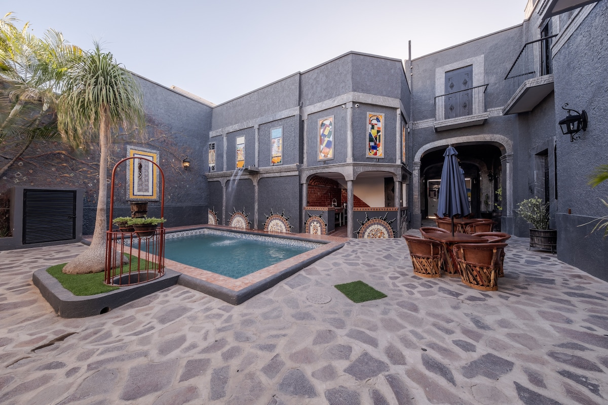 An outdoor area features a pool surrounded by stone paving, with lush palm trees adding greenery. Colorful stained glass accents the gray walls, while a few outdoor seating areas are visible, including large wooden chairs and an umbrella providing shade.