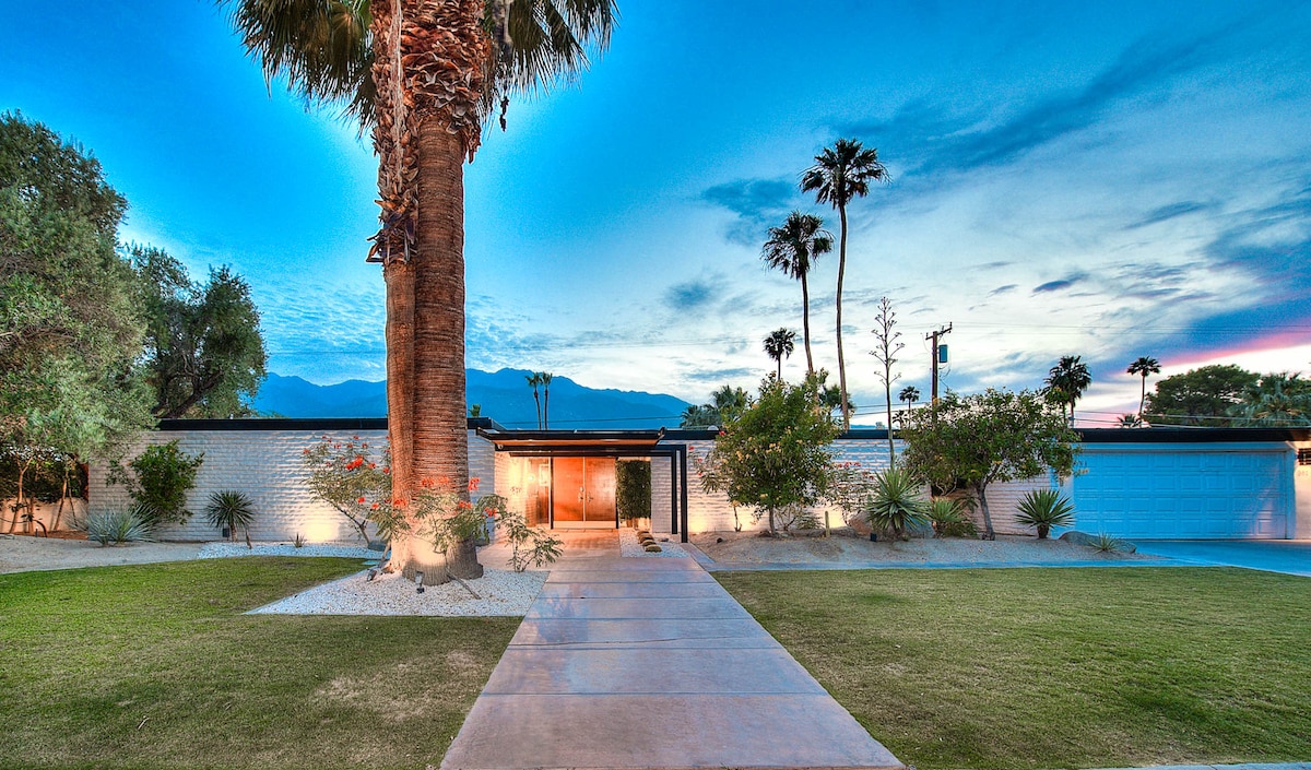 An inviting entrance is framed by palm trees against a twilight sky, showcasing a clean pathway leading to the entrance of the home. Lush landscaping and soft lighting create a serene atmosphere, highlighting the architectural lines of the facade.