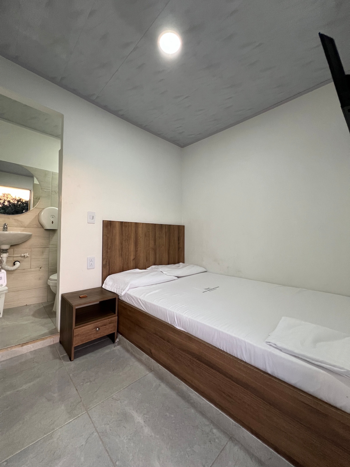 A compact room includes a single bed with a wooden headboard and a bedside table. A private bathroom is visible in the background, featuring modern fixtures. Natural light enters the room through a ceiling light, illuminating the neutral-colored walls.
