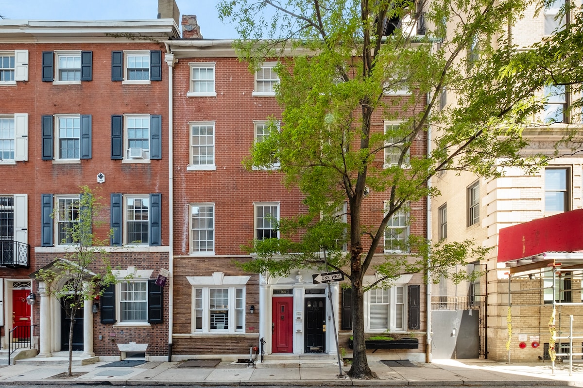 The exterior of the villa is showcased, featuring a classic red brick façade with white shutters and windows. A vibrant green tree is visible in front, providing a touch of nature to the urban setting. The entrance is marked by a bold red door.