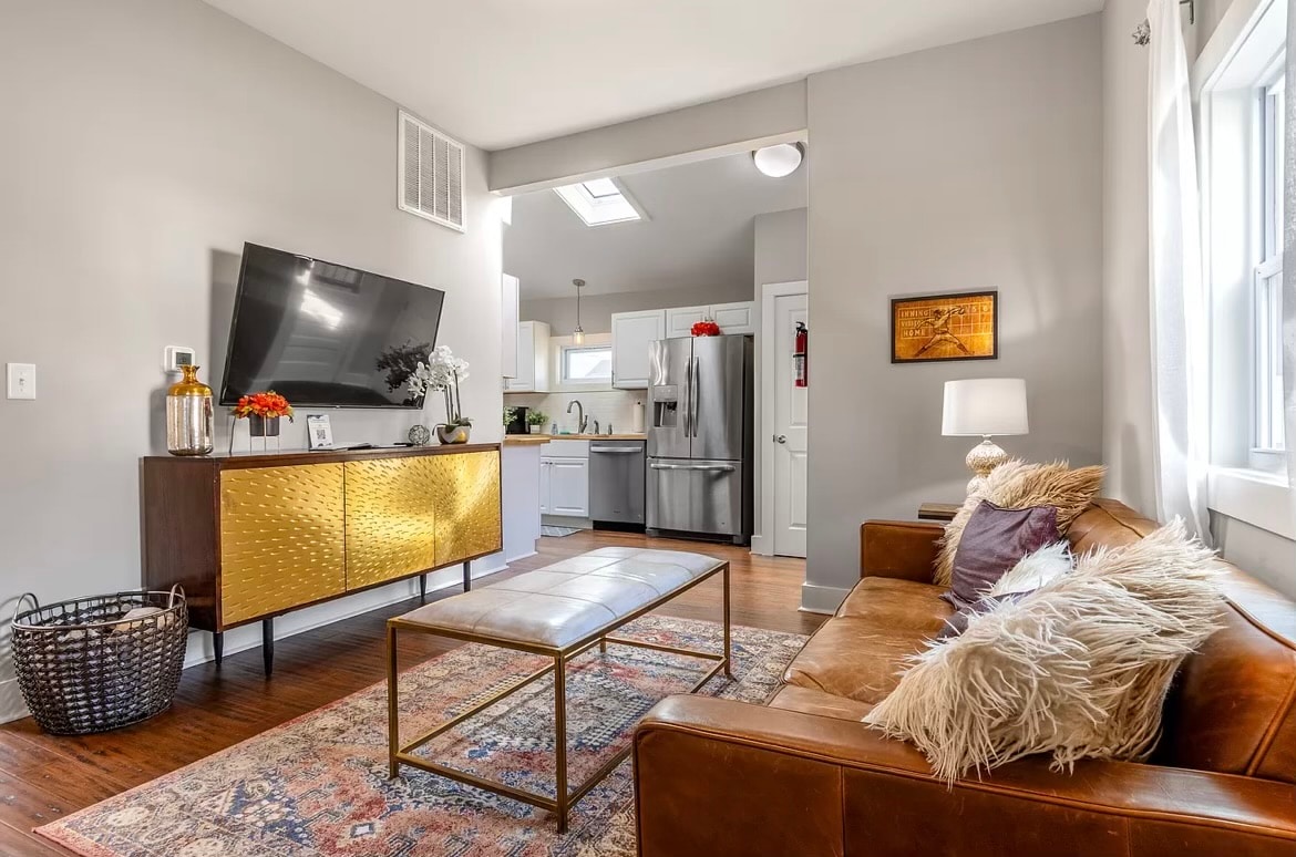 A modern living area features a stylish brown leather sofa complemented by decorative pillows. A mid-century coffee table sits at the center, with a textured surface. A flat-screen television is mounted on the wall above a sleek gold-accented cabinet, and natural light enters through nearby windows.