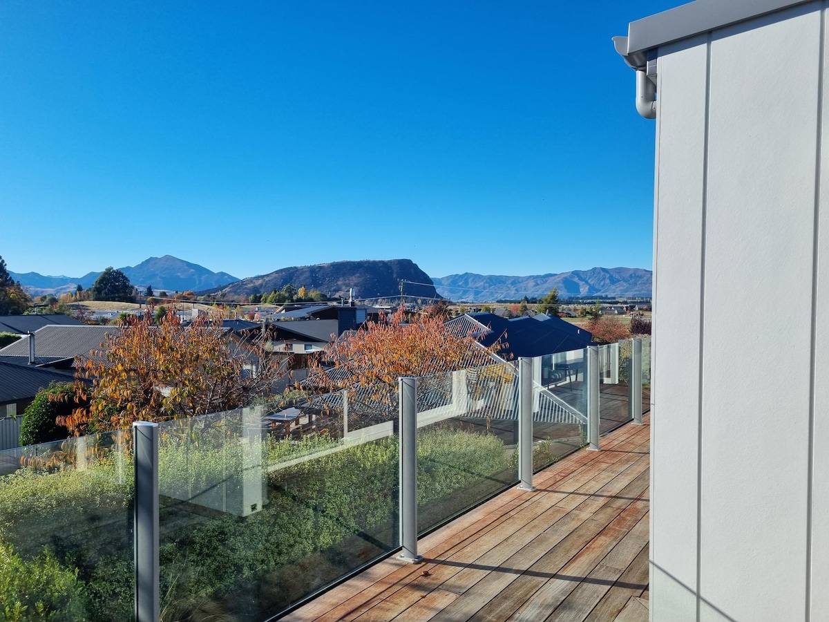 An outdoor terrace is showcased with a wooden deck and glass railings. The surrounding rooftops and trees are visible, with a backdrop of mountains under a clear blue sky, highlighting the natural beauty of the area.