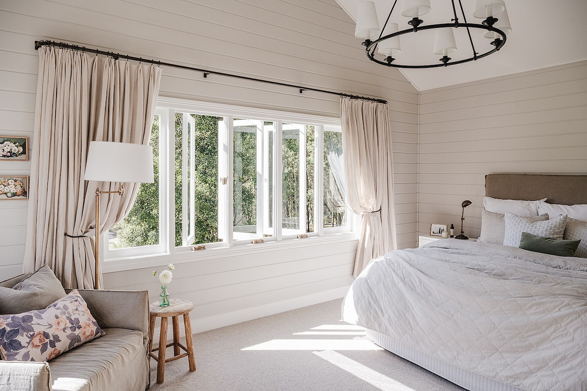 A well-lit bedroom is furnished with a spacious bed dressed in light linens. A small sitting area features a soft couch and a wooden stool. Large windows adorned with flowing curtains invite natural light, showcasing views of the greenery outside.