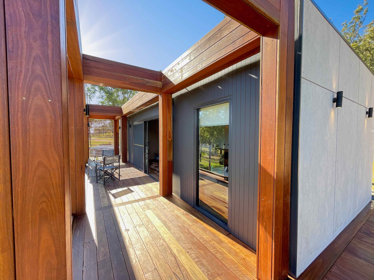 A spacious outdoor area is highlighted by wooden decking, providing a warm and natural setting. Large glass doors invite visibility to the interior, while modern outdoor seating arrangements are positioned for relaxation. Natural lighting floods the area, enhancing the connection with the surrounding environment.