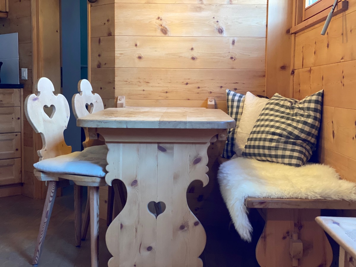 A charming dining area is visible, featuring a wooden table with a unique heart-shaped design and two matching chairs. Cushions with a checkered pattern and a fur throw are arranged on the bench seating, complemented by warm wooden walls that enhance the cozy ambiance.