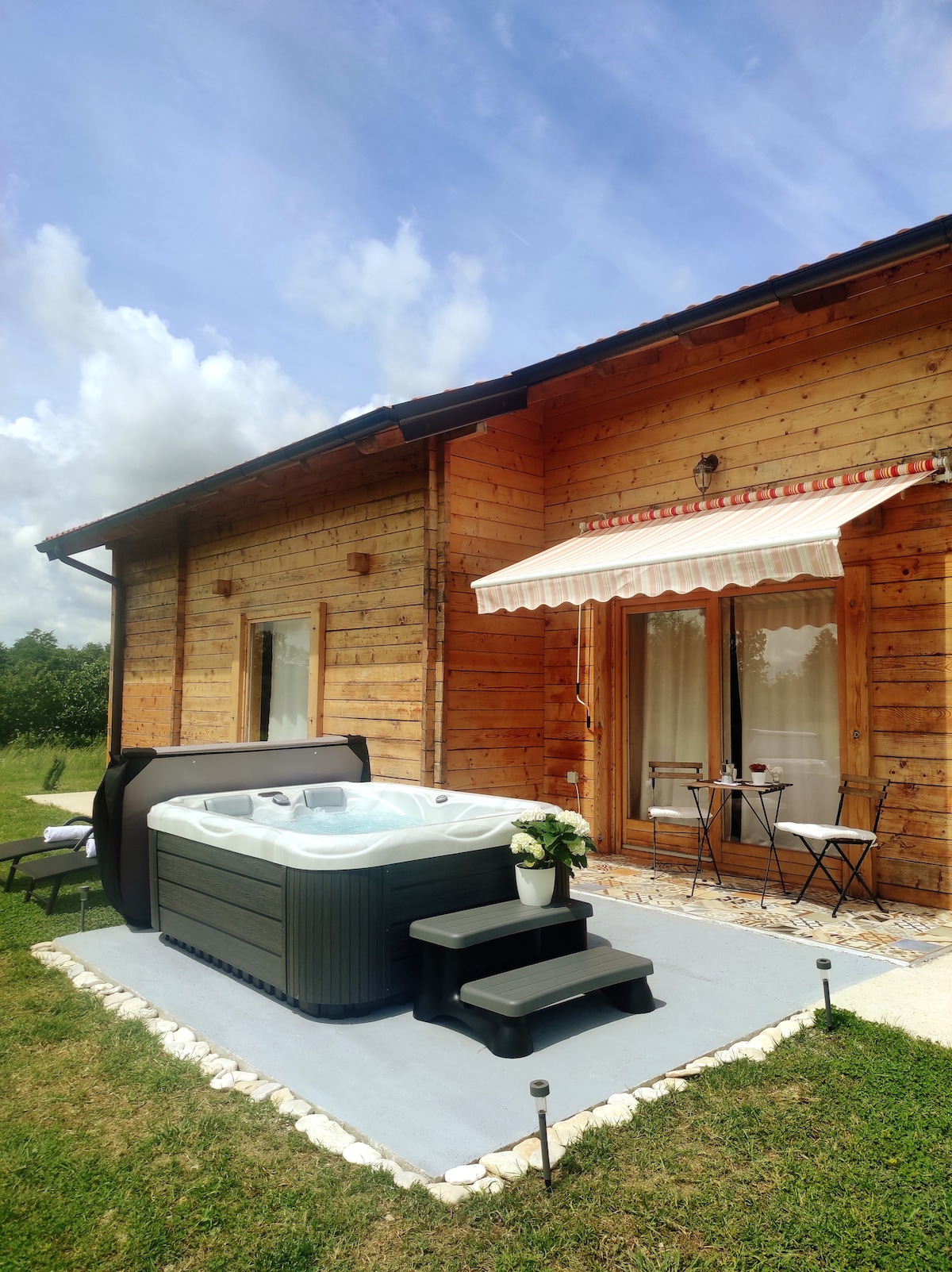 A secluded outdoor area features a private hot tub, surrounded by lush greenery. Nearby, a small table and chairs invite relaxation, with the wooden facade of the house providing a rustic backdrop. White stone borders frame the patio area, enhancing its tranquil setting.