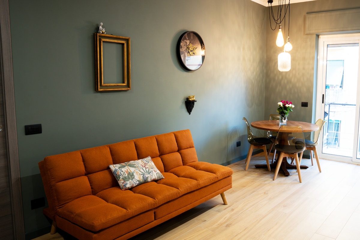 A modern living area is displayed with a bright orange sofa and a round dining table with clear chairs. Wall décor includes a circular mirror and a small flower vase, contributing to a relaxed and inviting ambiance. Natural light enters through the balcony doors.