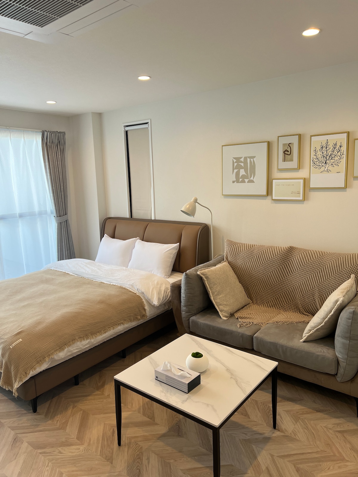 A modern bedroom features a spacious bed dressed in neutral linens, paired with a soft sofa adorned with textured throws. A small coffee table rests in the center, and natural light filters through a large window, enhancing the cozy atmosphere.