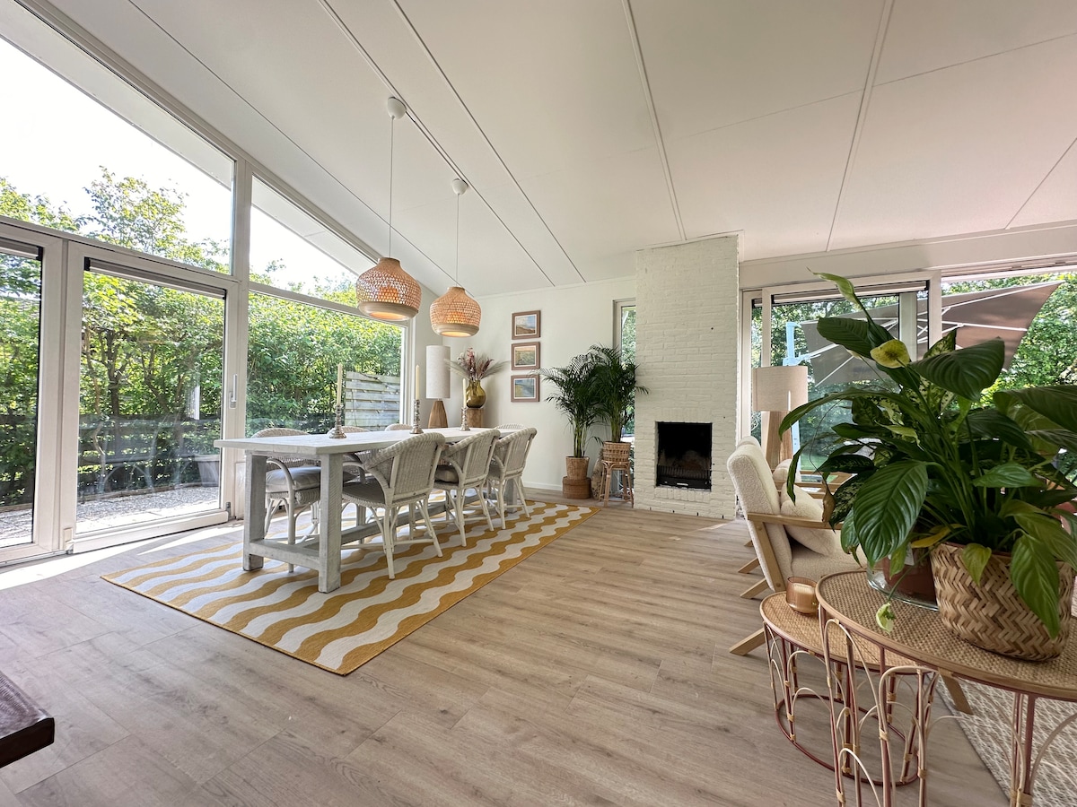 A spacious living area features large windows allowing natural light to fill the room. A dining table surrounded by light-colored chairs is positioned near a warm fireplace. Decorative plants and unique light fixtures add character to the inviting space.