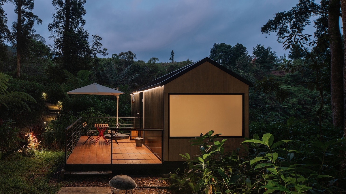 A modern cabin is nestled in a natural setting, featuring a wooden exterior and a cozy balcony. Two chairs with a small table are arranged under an umbrella, surrounded by lush greenery. Soft lighting highlights the structure during twilight.