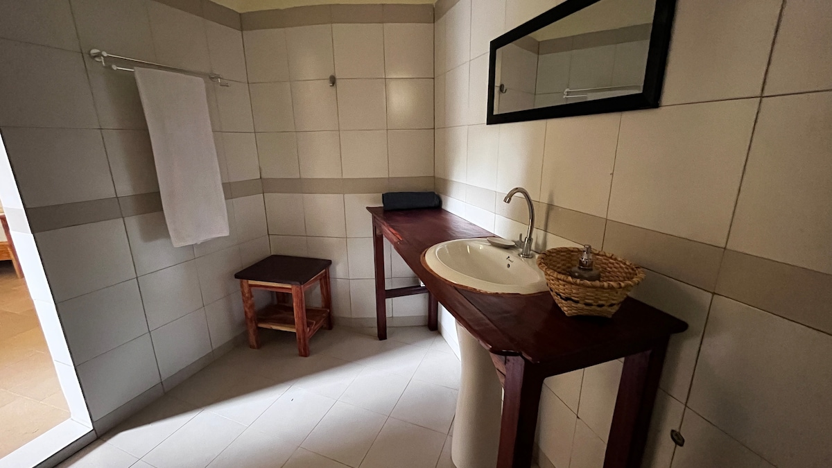 A modern bathroom features white tiled walls and a light-colored floor. A wooden vanity with a circular sink is positioned next to a rectangular mirror. A small wooden stool and a towel rack are also present, providing practical amenities.