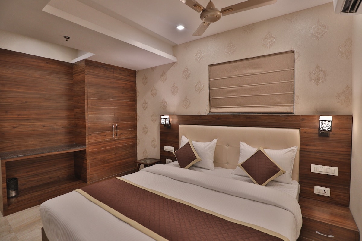 A well-appointed guest room features a spacious bed with a patterned bedspread. Wooden paneling lines the walls, complemented by subtle wall decor. Soft lighting is provided by stylish lamps on either side of the bed. A window with a fabric shade allows for natural light.