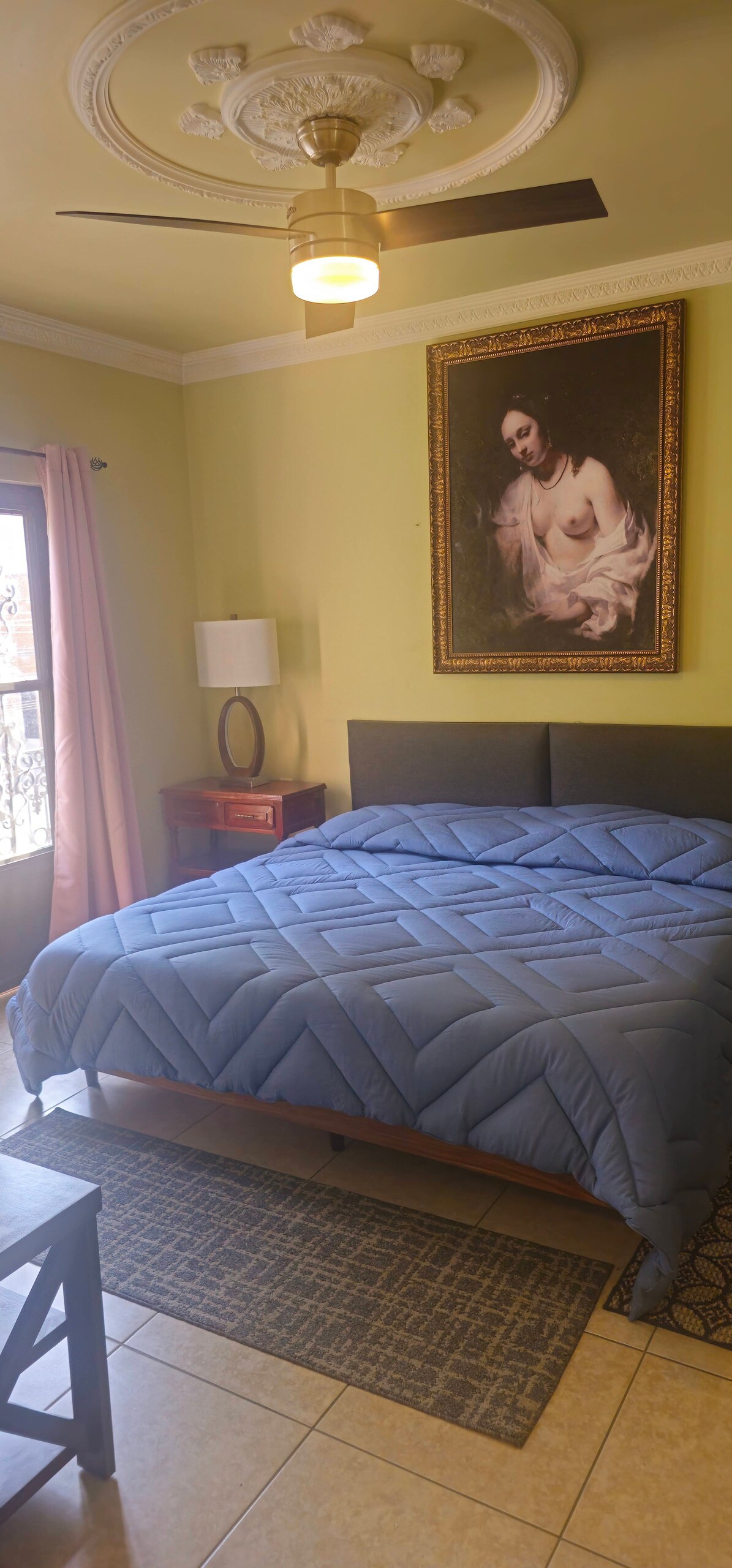 A sleeping area features a spacious bed covered with a blue quilt. Light green walls are complemented by a large framed artwork above the bed. A small side table with a lamp sits next to the bed, and a ceiling fan provides cooling. Natural light enters through the balcony doors.