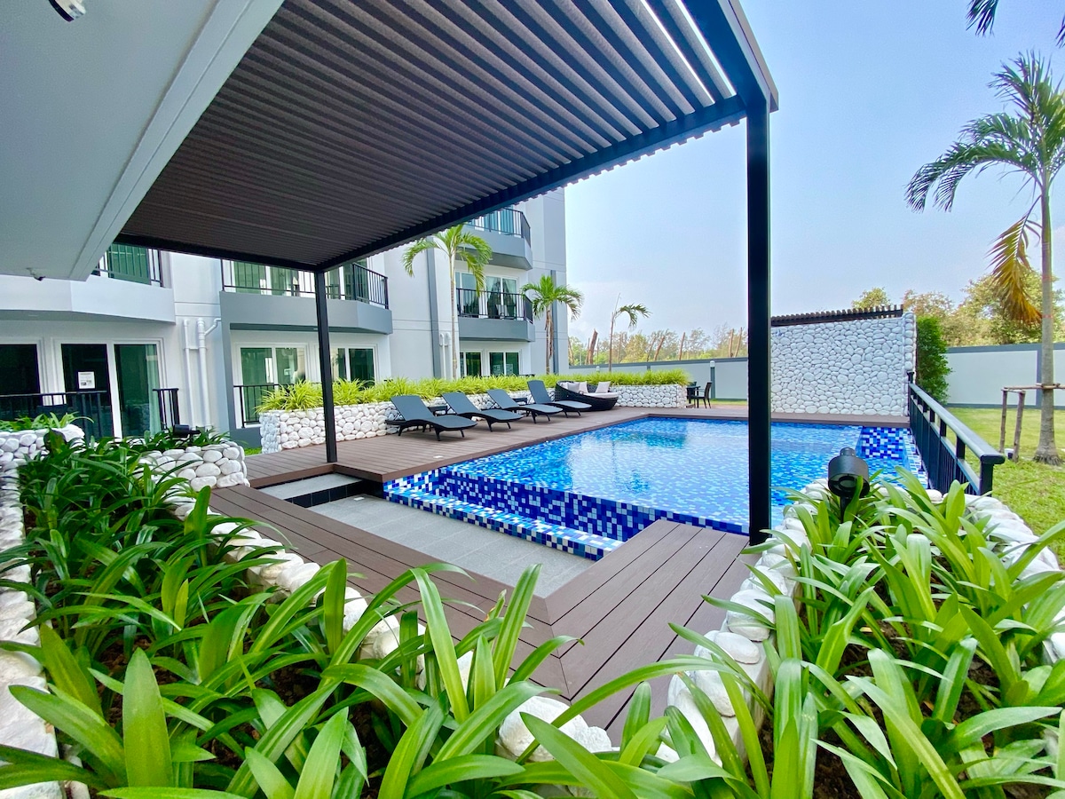 A swimming pool is surrounded by lush greenery and wooden decking, featuring several lounge chairs positioned for relaxation. The area is partially shaded, providing comfortable seating options near the water, with modern buildings visible in the background.