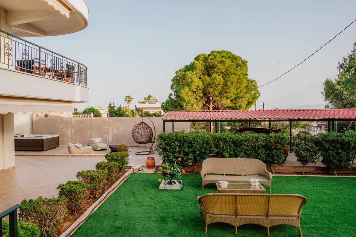 A spacious garden area is featured, featuring a lush green lawn and decorative plants. Comfortable seating arrangements are provided, including a pair of chairs and a small table. In the background, a Jacuzzi and a cozy swinging chair enhance the outdoor setting.