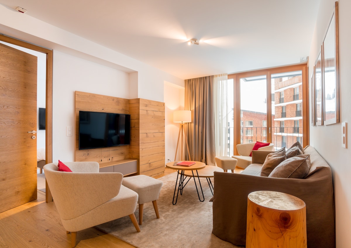 A cozy living area is presented, featuring a modern sofa and two armchairs, complemented by a wooden side table. A flat-screen television is mounted on a wooden panel. Natural light enters through large windows, adorned with soft curtains, illuminating the stylish decor.