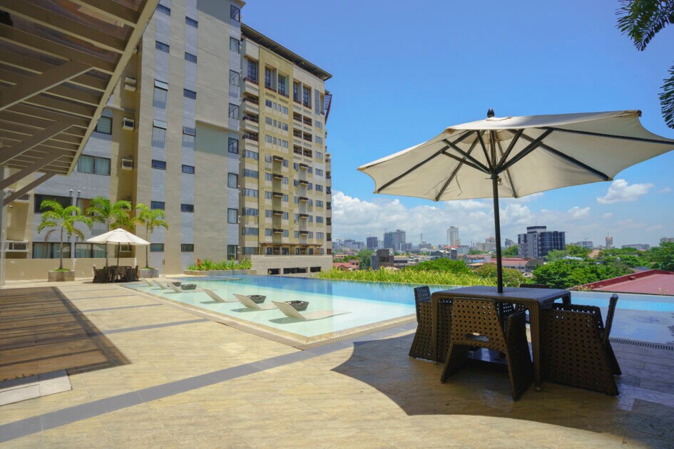 An outdoor area features a poolside deck with a patio table set beneath a large umbrella. The infinity pool reflects the skyline of Cebu City, framed by lush greenery and modern buildings in the distance under a clear blue sky.