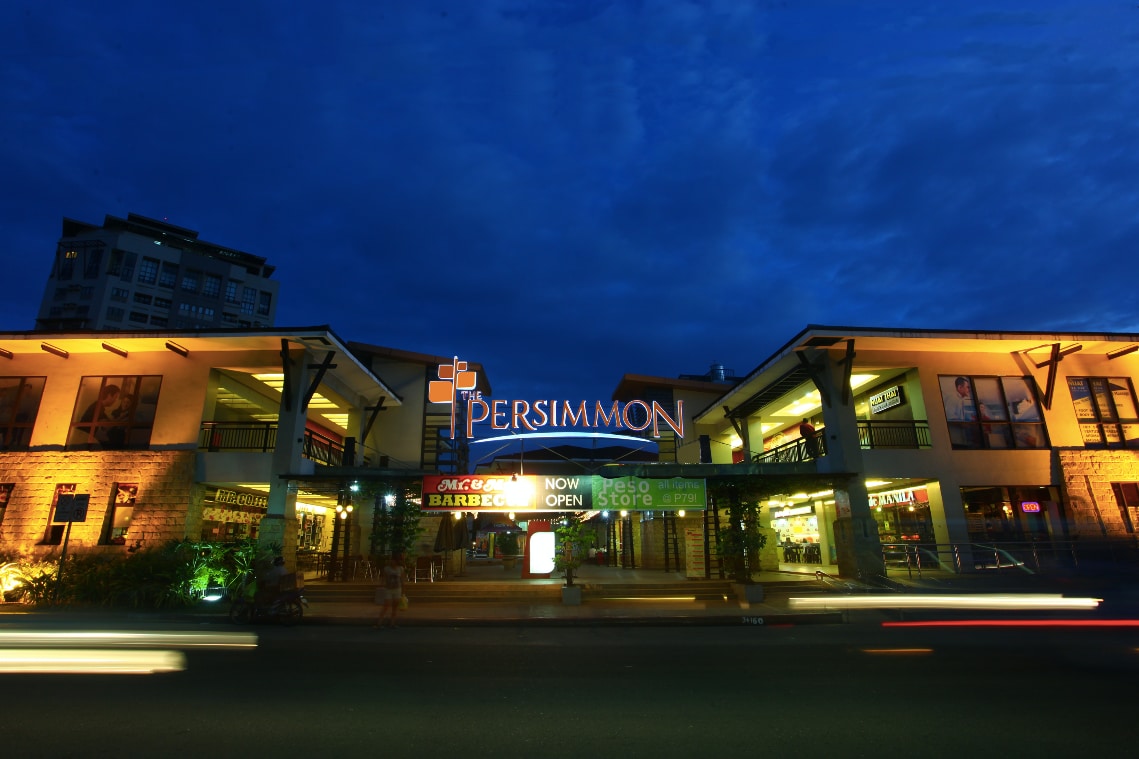 The exterior of the Persimmon retail complex is illuminated at dusk. The building features a mix of stone and modern design elements, with bright signage indicating various establishments. Trees and greenery line the entrance, adding to the urban environment.
