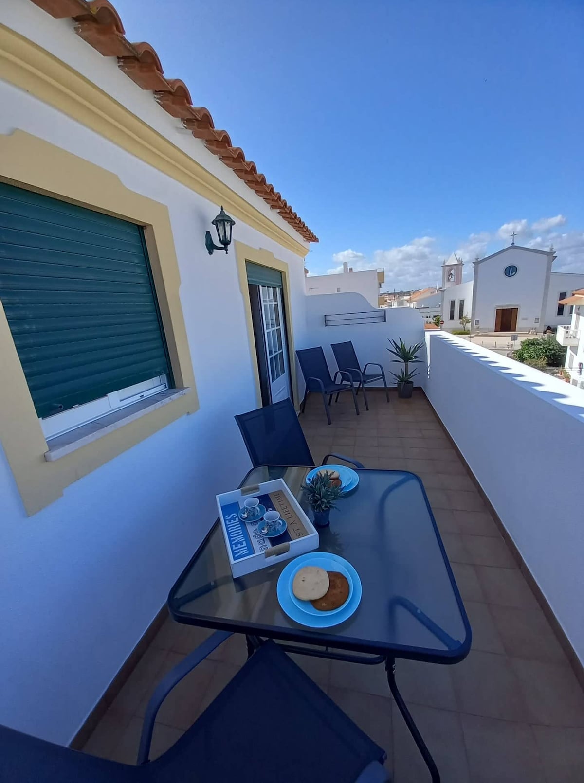 A balcony area is featured, furnished with a glass-top table and several chairs arranged for outdoor seating. A tray with drinks and snacks is placed on the table, while a small potted plant adds a touch of greenery. Scenic views of the surrounding area are visible.