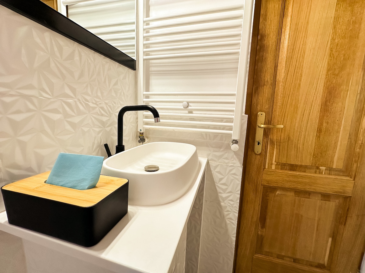 A modern bathroom features a sleek, circular sink atop a minimalist countertop. A stylish wooden box holds soft towels beside the sink. Decorative wall patterns add texture, while a wooden door provides access. An elegant faucet complements the contemporary design.