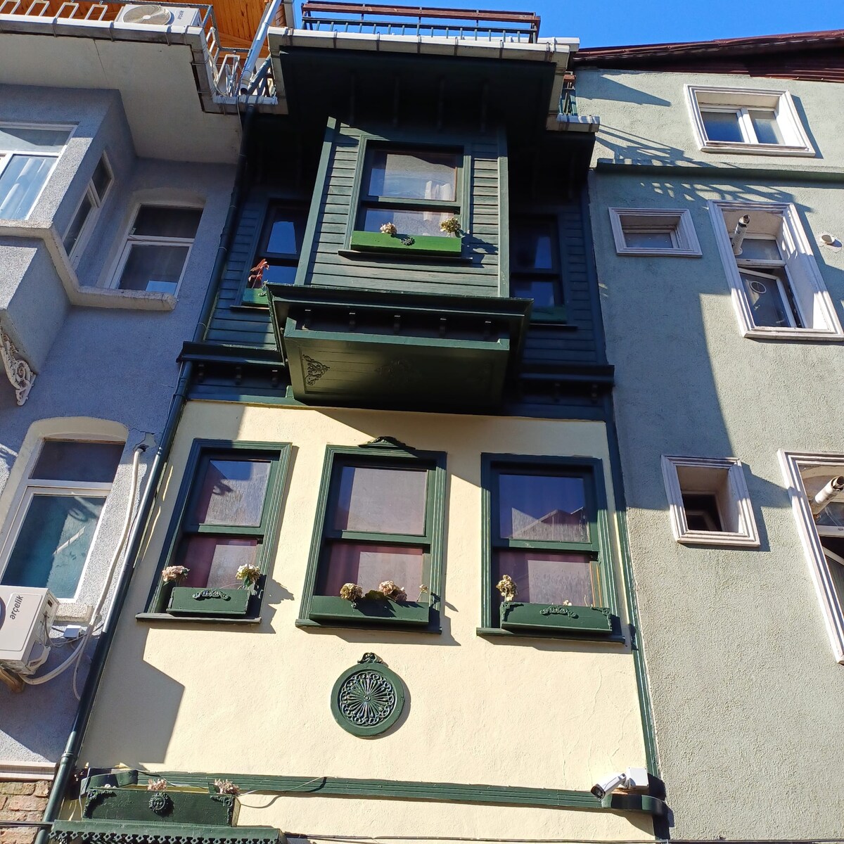 A charming multi-story building features an ornate green façade with decorative window frames. Window boxes adorned with flowers add vibrant touches to the exterior. The structure is flanked by two neighboring buildings, showcasing contrasting architectural styles and colors.