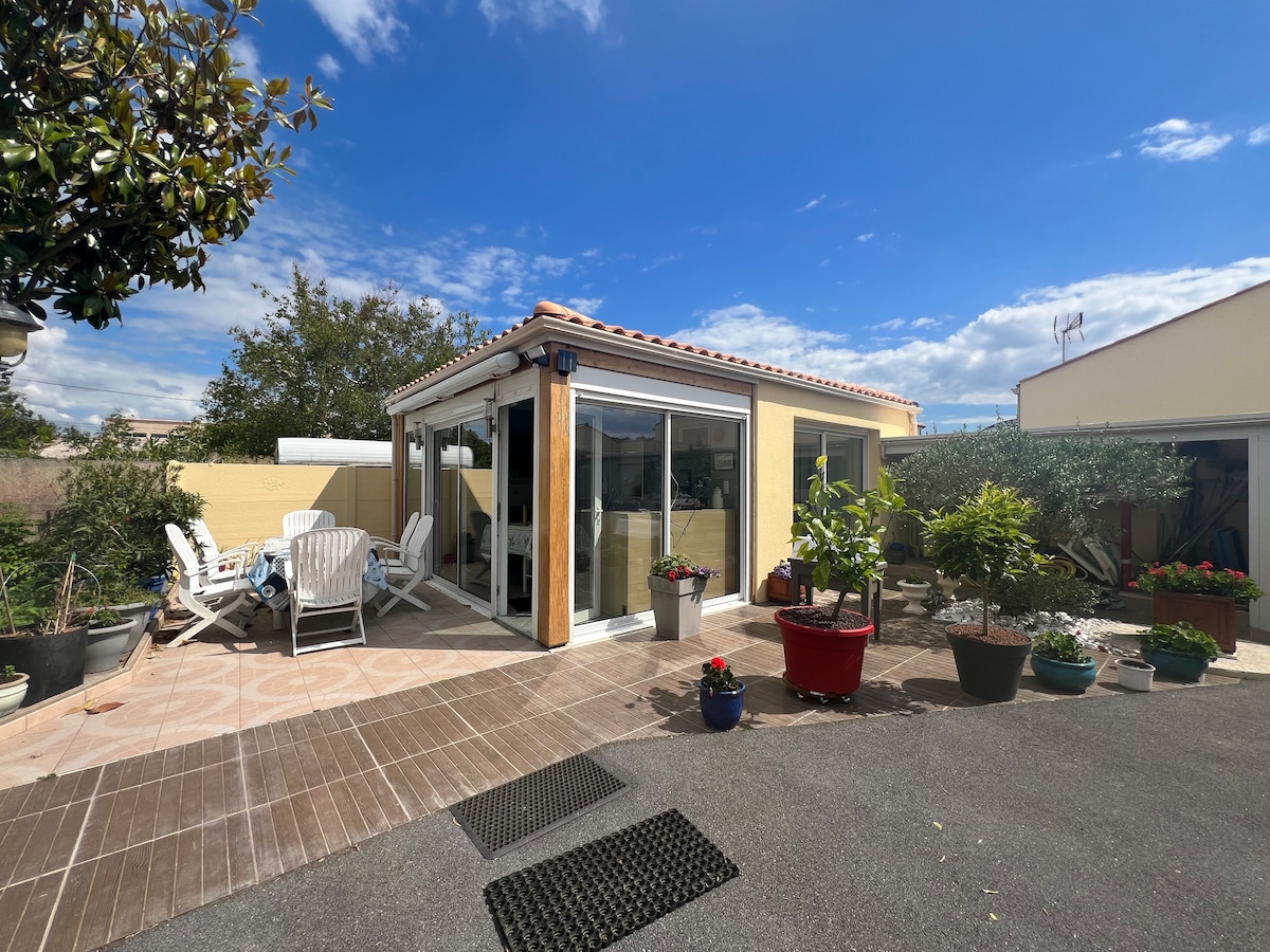 A bright outdoor area showcases a charming small house with large glass doors leading into a light-filled interior. Planters adorn the space, featuring a variety of greenery. Comfortable seating options are present, surrounded by well-tended plants under a clear blue sky.