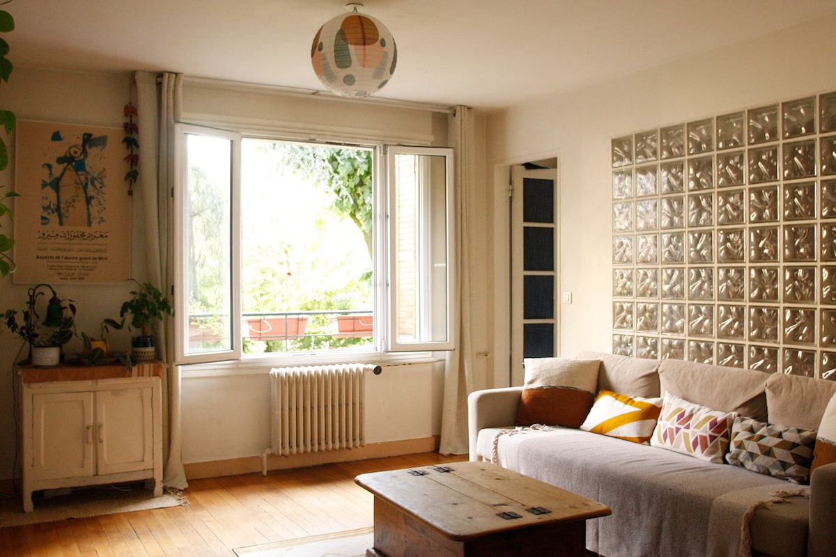 A spacious living area is illuminated by natural light filtering through large windows. A comfortable sofa adorned with colorful cushions is positioned in front of a wooden coffee table. An artistic poster hangs on the wall, while decorative plants are placed on a cabinet, enhancing the fresh atmosphere.