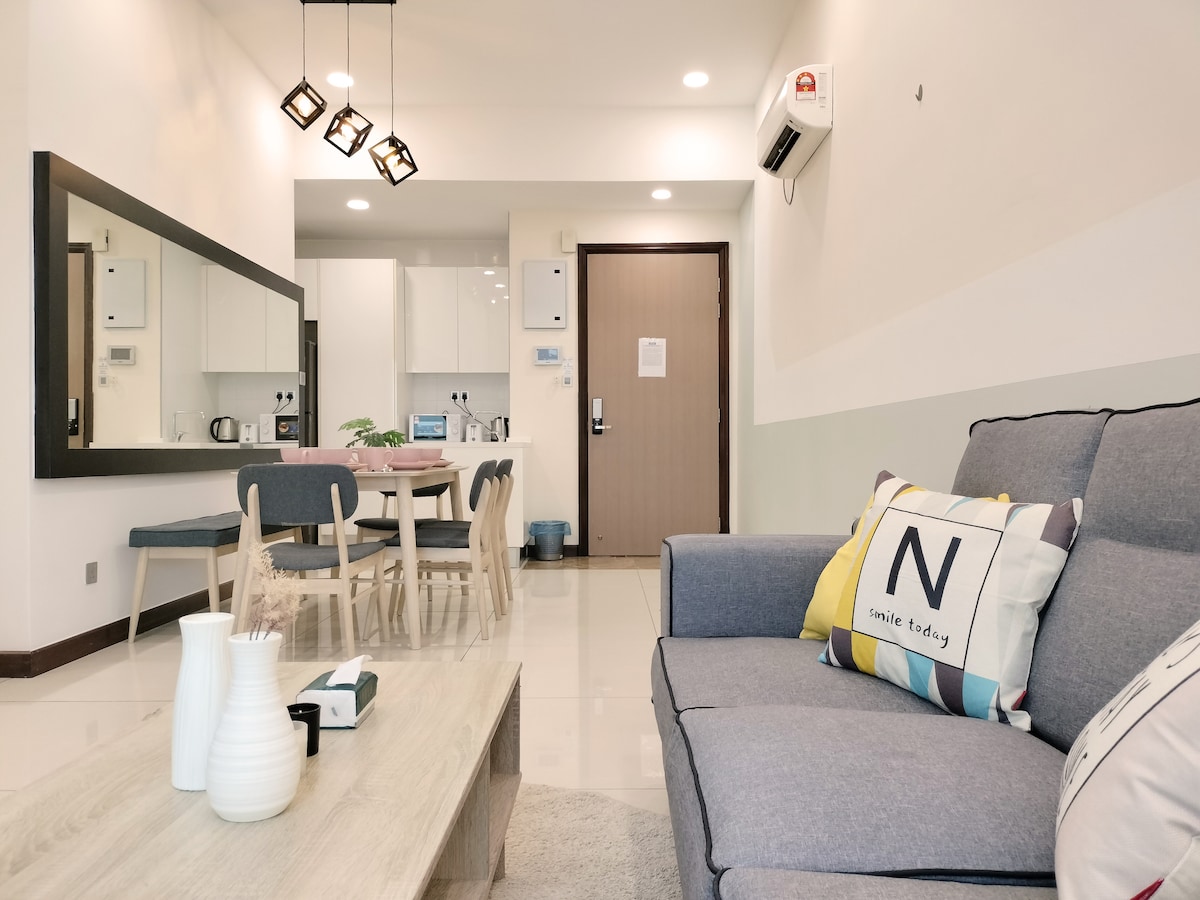 A modern living area features a comfortable grey sofa accompanied by decorative cushions. A wooden coffee table sits in front, and a dining area with a round table and chairs is visible in the background. Bright lighting highlights the open space, creating a welcoming atmosphere.