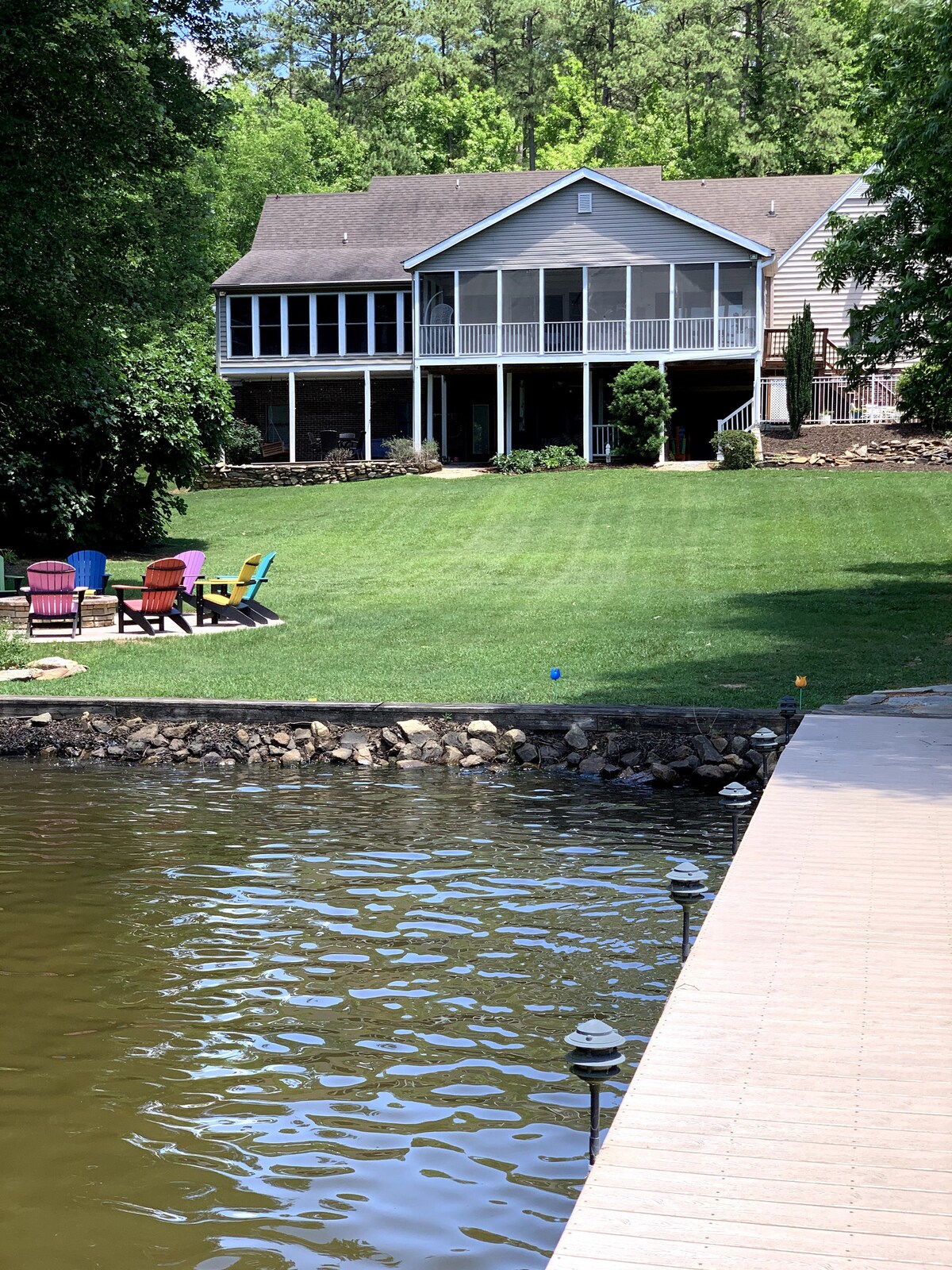 A spacious home is nestled among trees, featuring a screened-in porch and multiple outdoor seating areas. The well-maintained lawn extends toward a tranquil waterfront. A pathway leads to a dock, where lights illuminate the water, inviting relaxation and enjoyment.