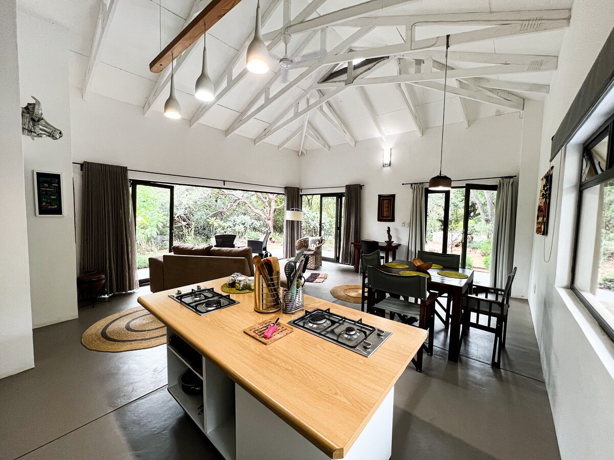 A spacious open-plan living area features polished concrete floors and high, white-beamed ceilings. A central kitchen island with a wooden countertop complements a cozy seating arrangement and a dining table. Large windows provide views of surrounding greenery, inviting natural light into the space.