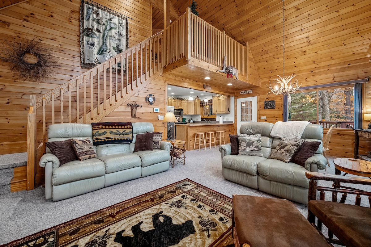 A spacious living area features comfortable seating with light-colored sofas arranged around a decorative rug. Wood paneling lines the walls, and a staircase leads to the upper level. A view of the kitchen area is visible in the background, illuminated by warm lighting.