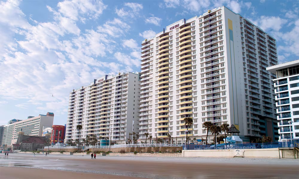 Two tall buildings line a pedestrian beach, showcasing modern architecture with balconies and large windows. A sandy beach stretches along the foreground, while a blue sky with scattered clouds frames the scene, enhancing the coastal setting.