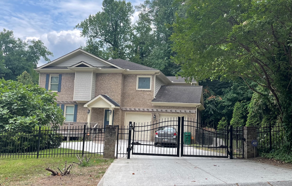 The exterior of a two-story home is framed by greenery, showcasing a brick facade and a gated entrance. A driveway leads to a spacious parking area. The house features multiple windows, and a car is parked in front.